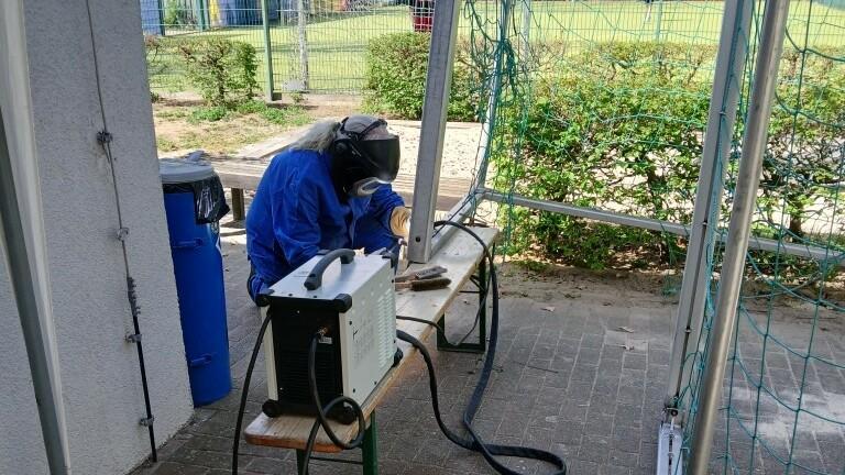 Arbeiter in blauer Schutzkleidung montiert Geräte an einem Tisch im Freien, umgeben von einer Sportanlage.