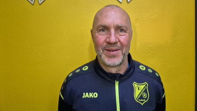 Porträt eines lächelnden Mannes in Sportjacke vor gelber Wand mit Fußball-Trophäen im Hintergrund.