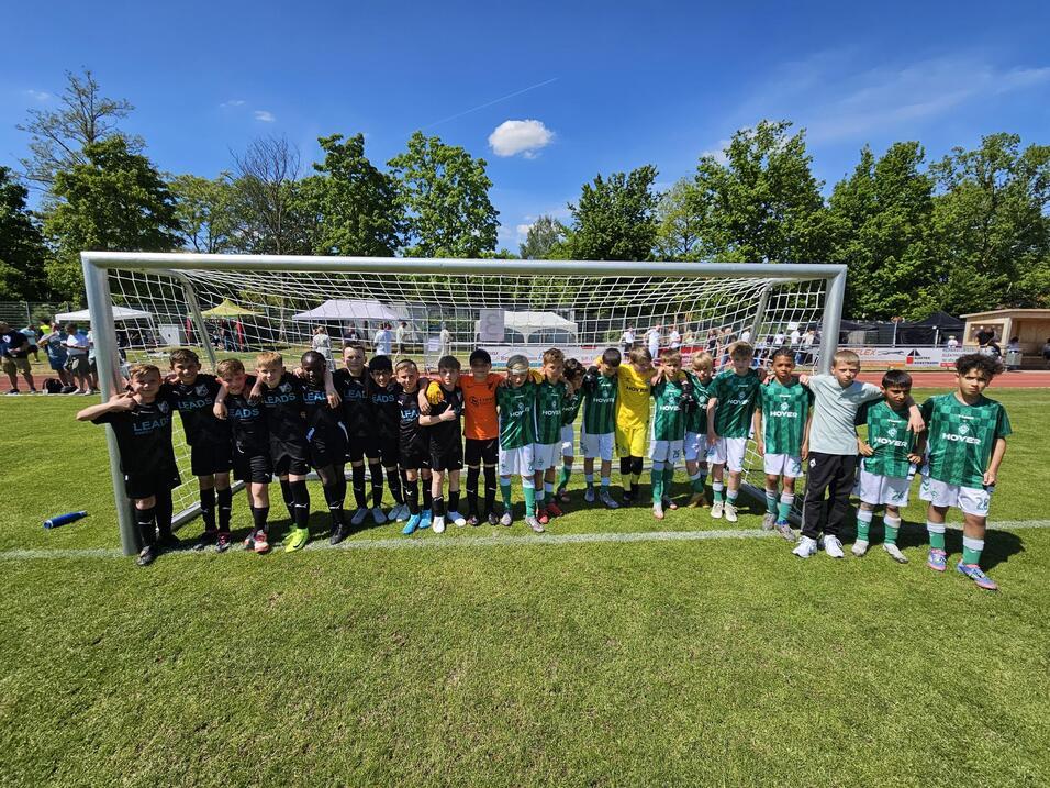 Zwei Fußballmannschaften stehen nebeneinander vor einem Tor auf einem grünen Spielfeld, umgeben von Bäumen und Zuschauern.