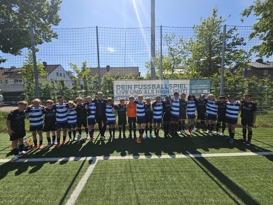 GruppennFoto von Jungen in Fußballtrikots auf einem Kunstrasenplatz, umgeben von einem netz eingezäunten Bereich.