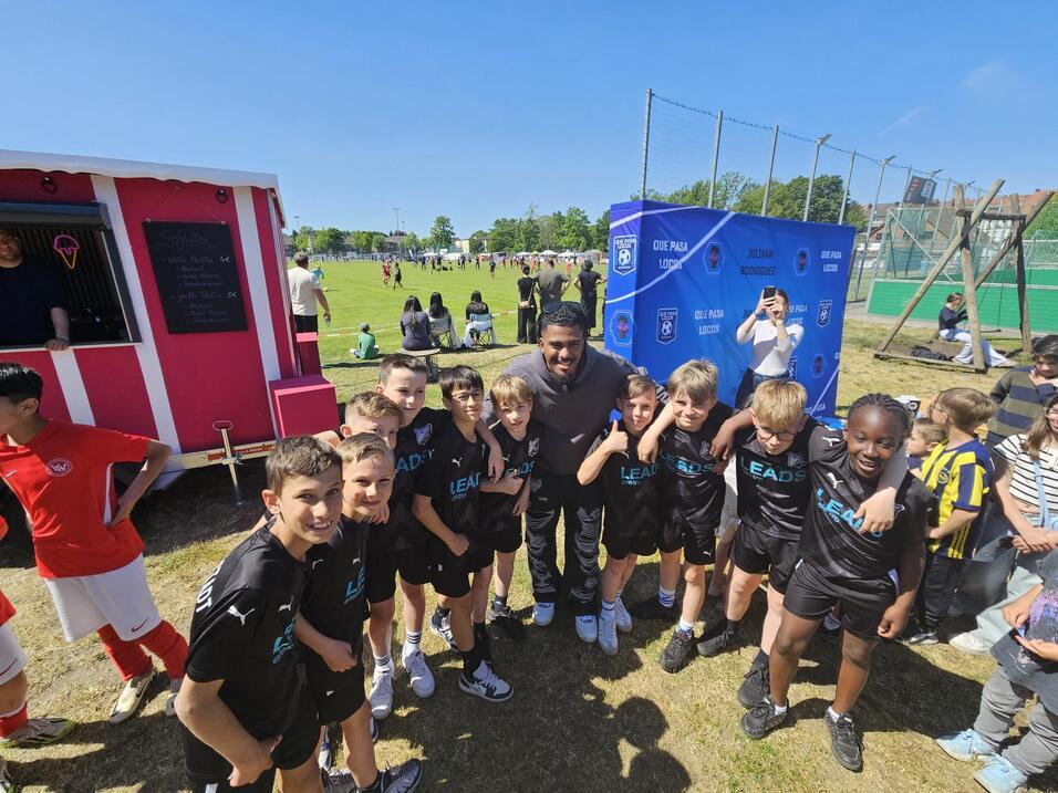 Gruppe von Kindern in Sportbekleidung posiert lächelnd mit einem Erwachsenen vor einem Fußballspiel im Freien.