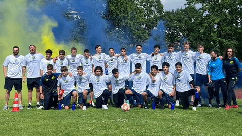 Gruppenfoto einer Fußballmannschaft in weißen Trikots, umgeben von buntem Rauch, auf einer Wiese.