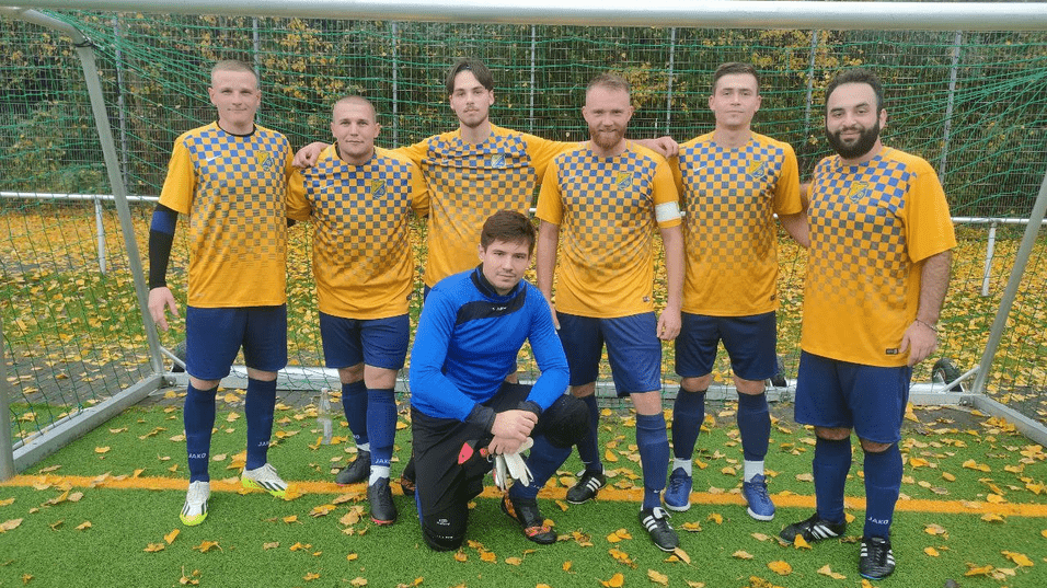 Sechsköpfige Fußballmannschaft in gelben Trikots mit Karomuster steht vor dem Tor auf einem mit Laub bedeckten Platz.