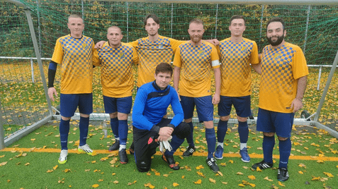 Sechsköpfige Fußballmannschaft in gelben Trikots mit Karomuster steht vor dem Tor auf einem mit Laub bedeckten Platz.