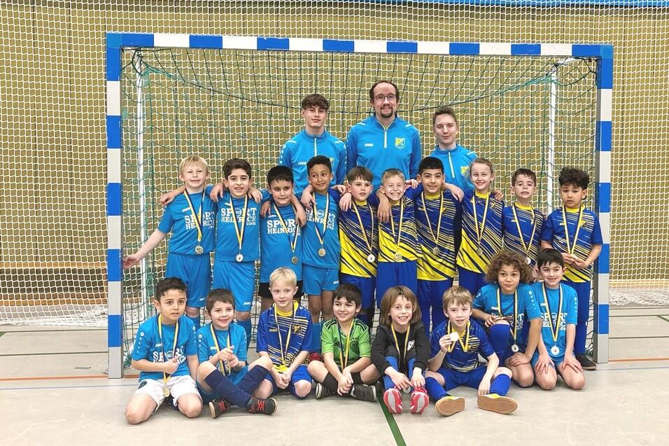 Gruppenkfoto von jungen Fu&szlig;ballspielern in Sporttrikots mit Medaillen, vor einem Tor in einer Sporthalle.