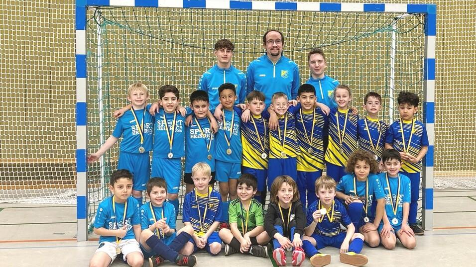 Gruppenkfoto von jungen Fußballspielern in Sporttrikots mit Medaillen, vor einem Tor in einer Sporthalle.