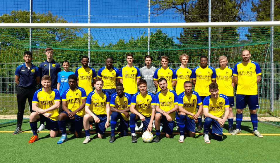 Gruppenfoto einer Fußballmannschaft in gelben Trikots auf einem Platz mit Fußballtor und Netz im Hintergrund.