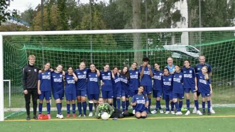 Mannschaftsfoto eines Mädchenfußballteams in blauen Trikots vor einem Fußballtor auf einem Kunstrasenplatz.