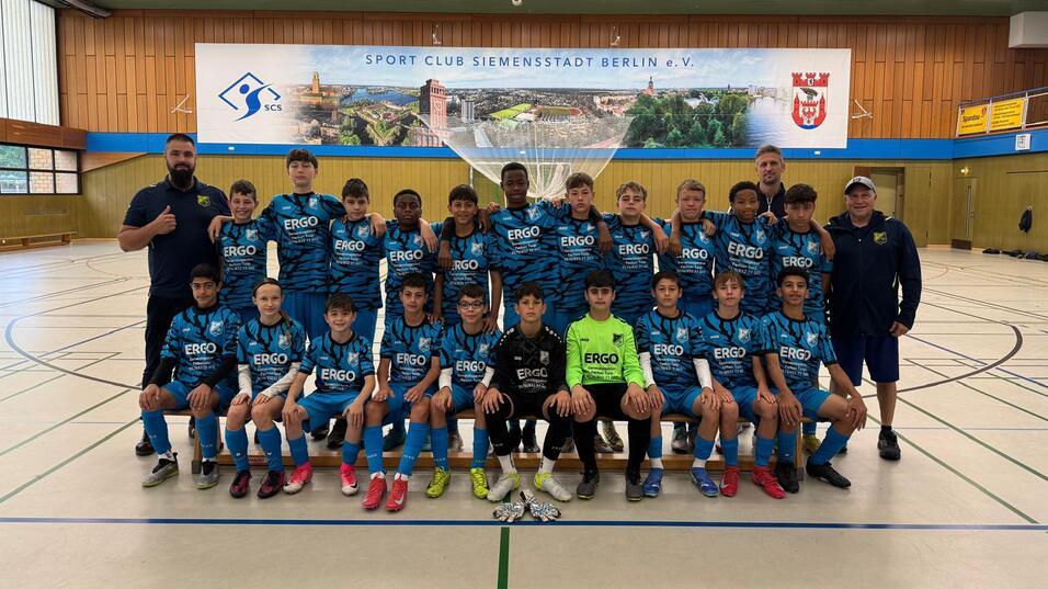 Jugendfußballteam in blauen Trikots posiert mit Trainern in einer Sporthalle von Berlin. Banner des Sportvereins im Hintergrund.