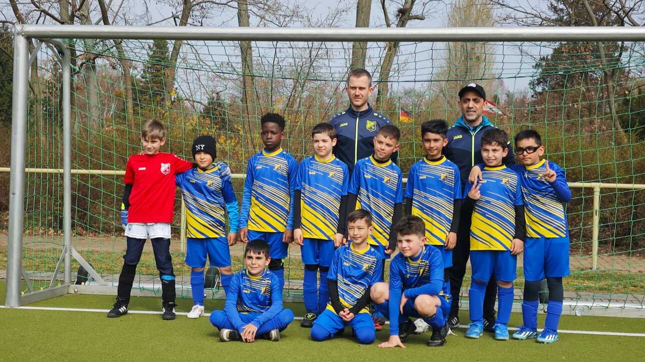 Gruppenfoto einer Fußballmannschaft mit neun Spielern und zwei Trainern vor einem Tor auf einem Sportplatz.