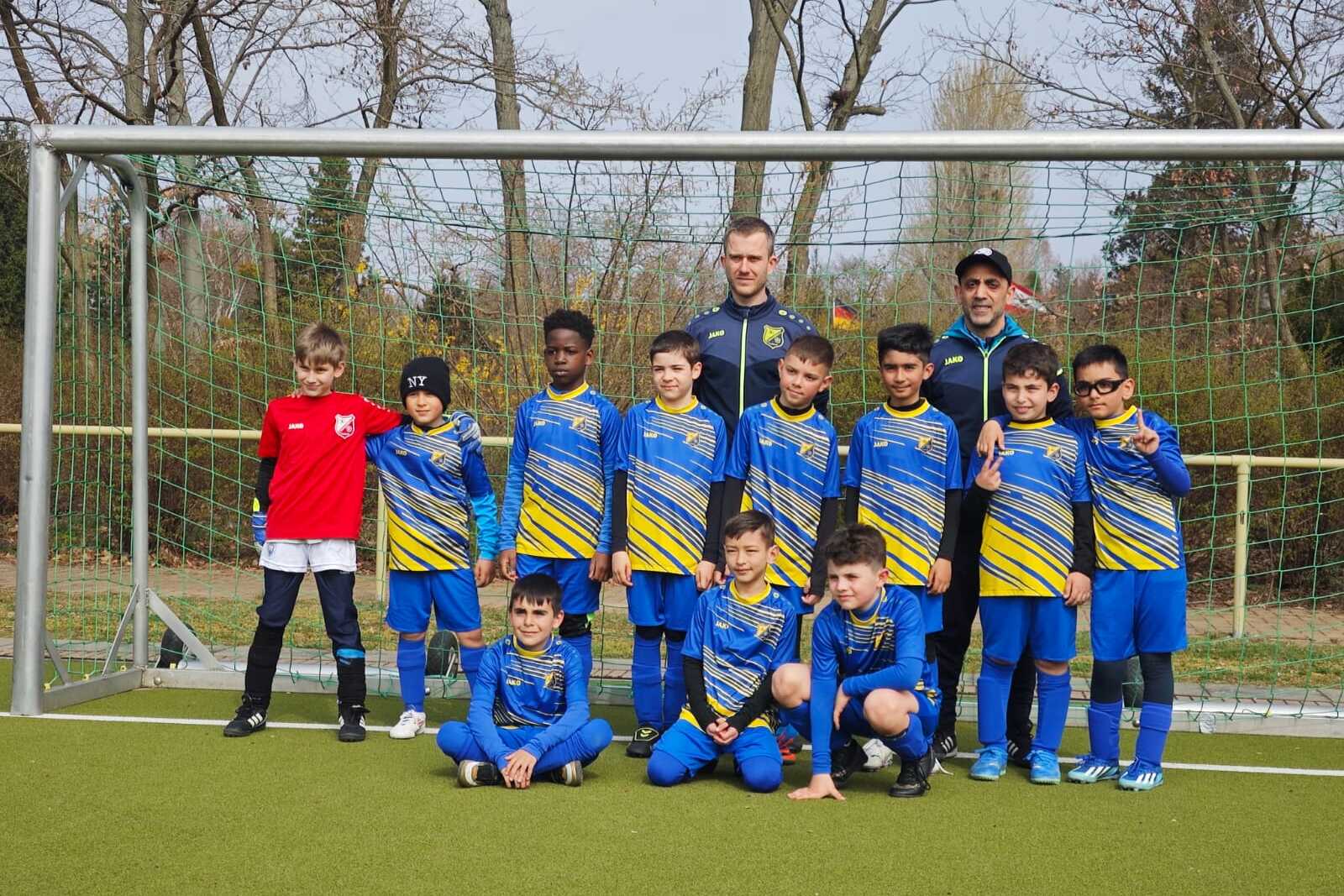 Gruppenfoto einer Fu&szlig;ballmannschaft mit neun Spielern und zwei Trainern vor einem Tor auf einem Sportplatz.