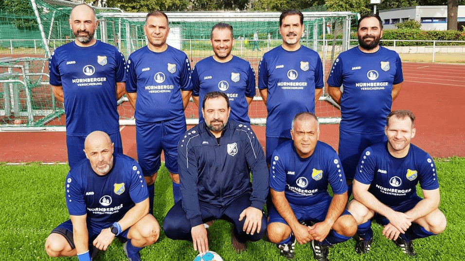 Gruppenfoto von acht Fußballspielern in blauen Trikots mit einem Ball auf einem Sportplatz vor einem Tor.