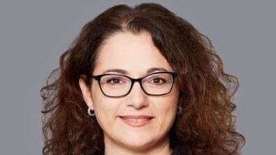 Porträt einer Frau mit lockigem Haar und Brille, die ein schwarzes Hemd trägt, vor grauem Hintergrund.