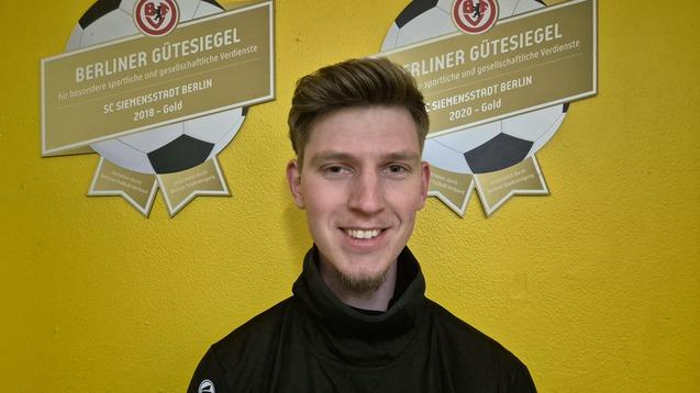 Junger Mann mit blondem Haar und Sportjacke lächelt vor gelbem Hintergrund mit zwei „Berliner Gütesiegel“-Auszeichnungen.