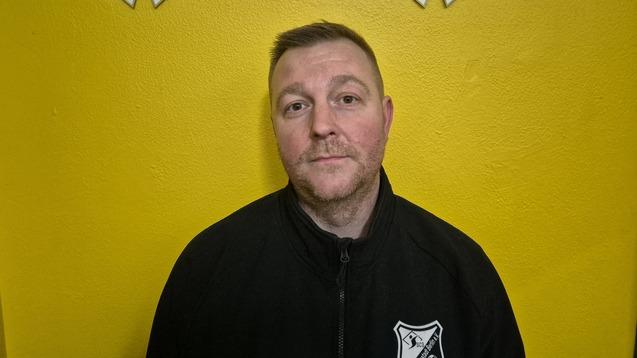 Porträt eines Mannes mit kurzem Haar, der in einem schwarzen Sweatshirt vor einer gelben Wand steht.