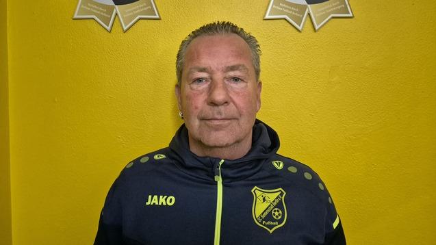 Mann mit kurzem Haar, trägt eine Trainingsjacke, vor gelber Wand und zwei goldenen Sportauszeichnungen.