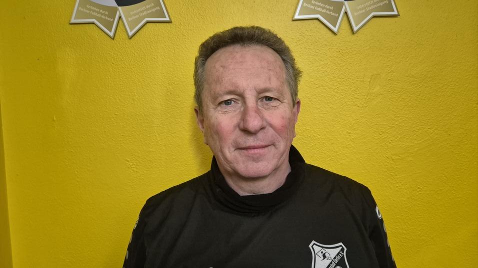 Mann in schwarzem Trainingsanzug mit Vereinslogo vor einer gelben Wand, lächelt in die Kamera.