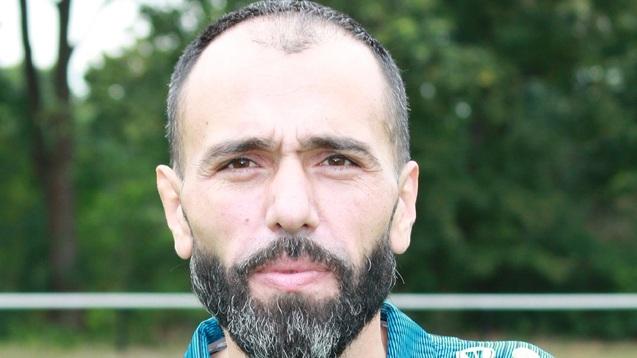 Porträt eines Mannes mit Bart in Sportkleidung, Hintergrund mit verschwommenem Fußballplatz.