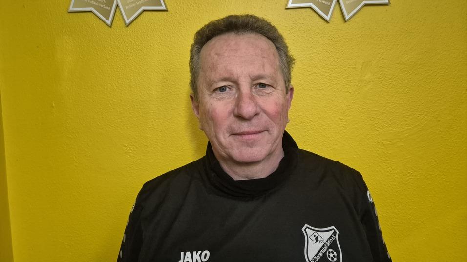 Mann in schwarzem Trainingsanzug mit Vereinslogo vor einer gelben Wand, lächelt in die Kamera.