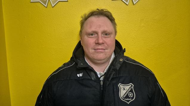 Mann im schwarzen Sportjackett mit Vereinslogo vor einer gelben Wand, zwei Fußball-Trophäen über ihm.