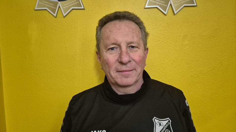 Mann in schwarzem Trainingsanzug mit Vereinslogo vor einer gelben Wand, lächelt in die Kamera.