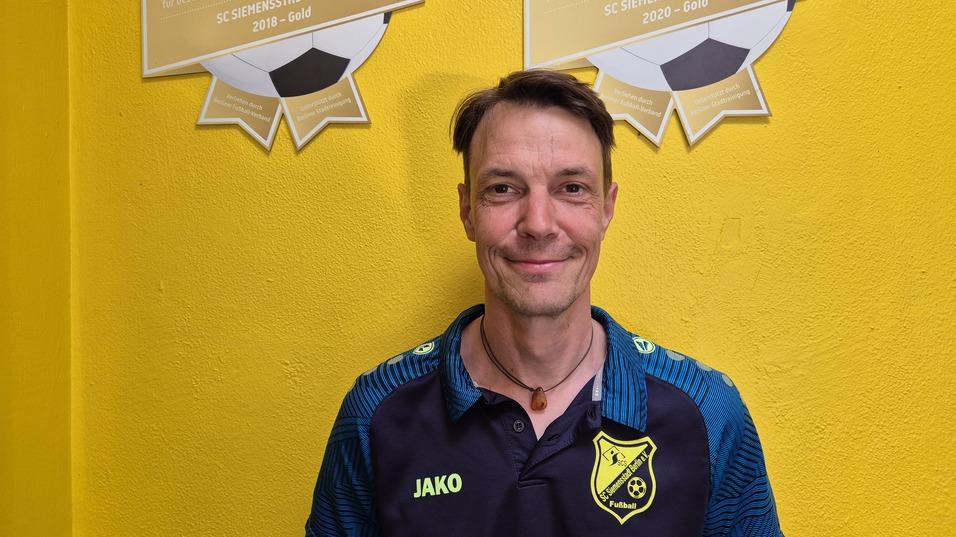 Mann mit sportlichem Oberteil steht vor einer gelben Wand, hinter ihm hängen Fußball-Ehrentafeln.