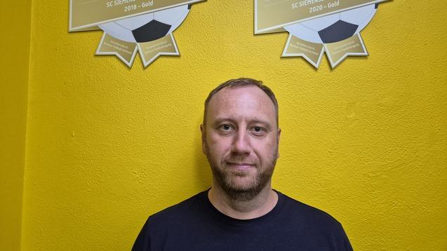 Mann steht vor einer gelben Wand mit zwei Berliner Gütesiegeln des SC Siemensstadt Berlin in Gold für 2018 und 2020.