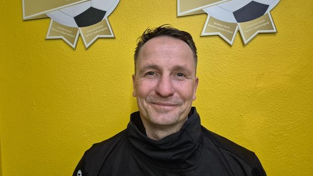 Mann in schwarzer Jacke l&auml;chelt vor einer gelben Wand mit zwei Fu&szlig;ballauszeichnungen im Hintergrund.