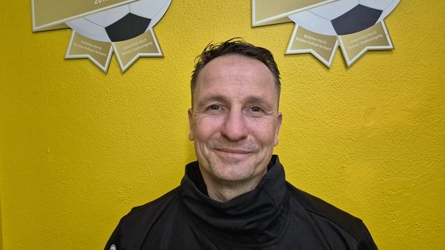 Mann in schwarzer Jacke lächelt vor einer gelben Wand mit zwei Fußballauszeichnungen im Hintergrund.