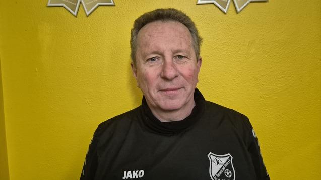 Mann in schwarzem Trainingsanzug mit Vereinslogo vor einer gelben Wand, lächelt in die Kamera.