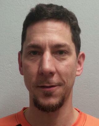 Porträt eines Mannes mit kurzem, dunklem Haar und Spitzbart, wearing orangeem Shirt vor grauem Hintergrund.