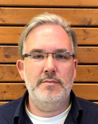 Mann mit Brille und Bart vor einer Holzlattenwand, Blick direkt in die Kamera gerichtet.