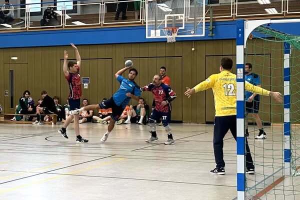 Handballspiel in der Halle: Spieler in blauer Trikots f&uuml;hren einen Sprungwurf aus, w&auml;hrend Gegner versuchen zu blocken.