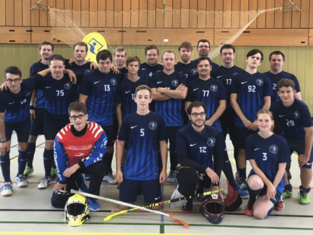 Gruppenfoto von 18 Personen in blauen Teamtrikots, mit Schlägern und Torwartausrüstung, in einer Sporthalle.