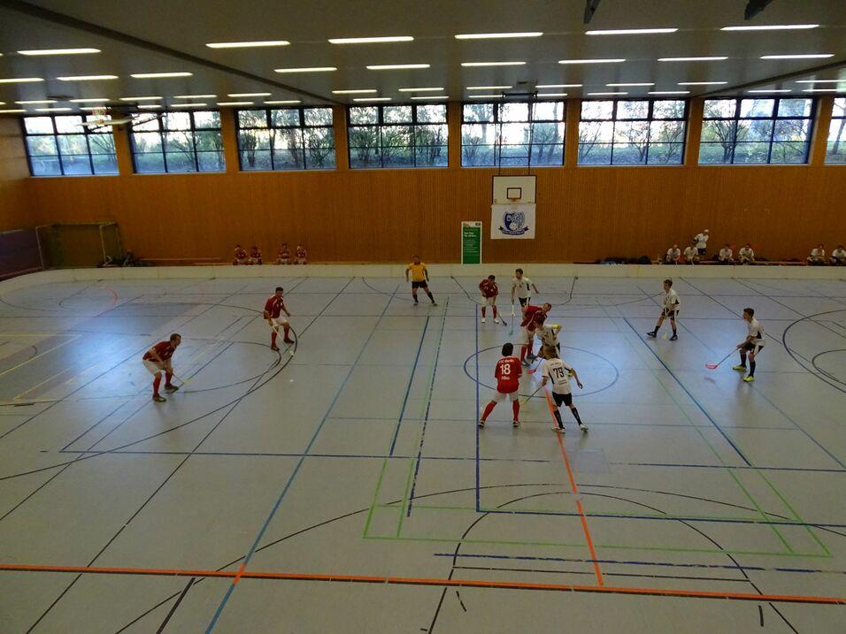 Zwei Teams in roten und weißen Trikots spielen in einer Sporthalle Hockey auf einem linienmarkierten Boden.