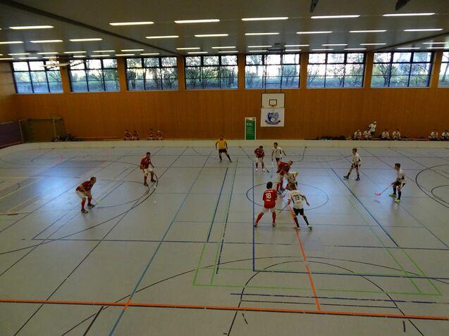 Zwei Teams in roten und weißen Trikots spielen in einer Sporthalle Hockey auf einem linienmarkierten Boden.