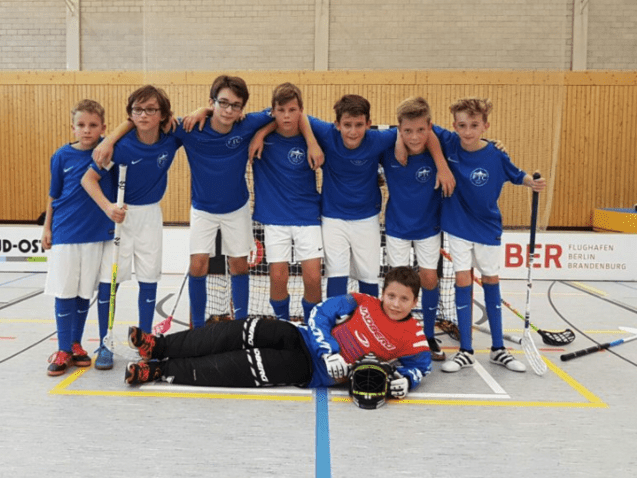 Gruppenfoto von acht Jungen in blauen Trikots mit Schlägern, stehen und sitzen vor einem Tor in einer Sporthalle.