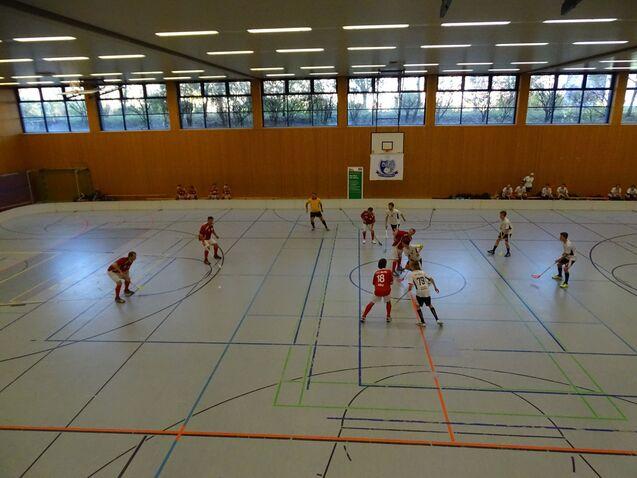 Zwei Teams in roten und wei&szlig;en Trikots spielen in einer Sporthalle Hockey auf einem linienmarkierten Boden.