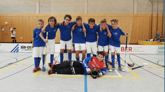 Gruppenfoto von acht Jungen in blauen Trikots mit Schl&auml;gern, stehen und sitzen vor einem Tor in einer Sporthalle.