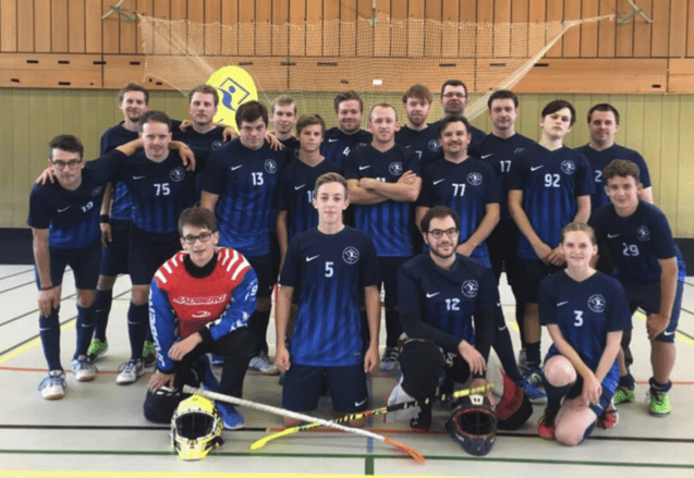 Gruppenfoto von 18 Personen in blauen Teamtrikots, mit Schlägern und Torwartausrüstung, in einer Sporthalle.