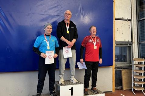 Drei Medaillengewinner stehen auf einem Podest und halten Urkunden in einer Sporthalle.