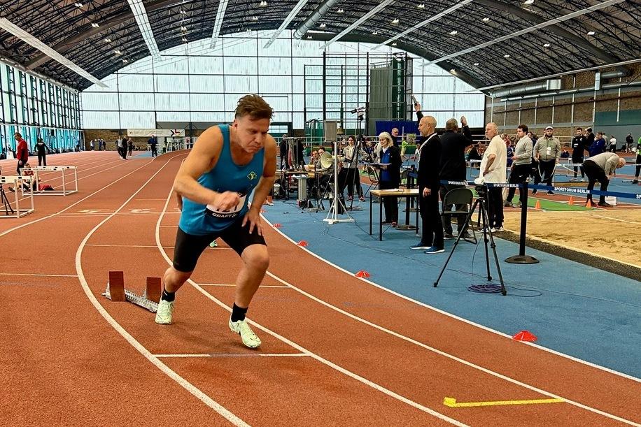 Ein Athlet startet sprintend auf einer Leichtathletikbahn in einer Sporthalle. Zuschauer und Geräte im Hintergrund.