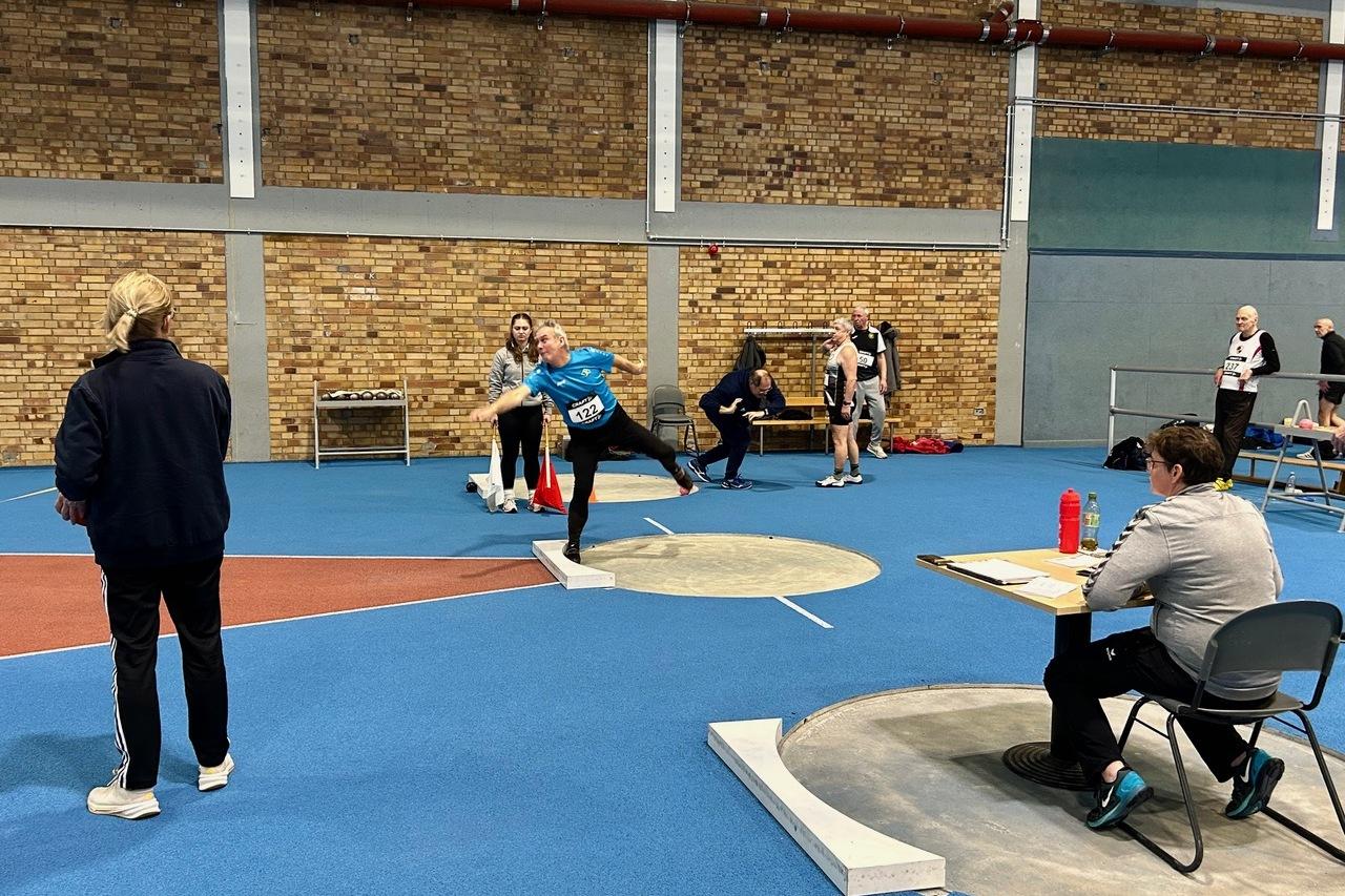 Athlet wirft in blauer Sportkleidung eine Kugel in einer Sporthalle, während Zuschauer und Kampfrichter beobachten.