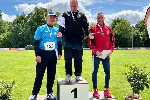 Drei Medaillengewinner stehen auf dem Siegertreppchen im Freien; der Erste trägt eine Medaille und hält ein Zertifikat.