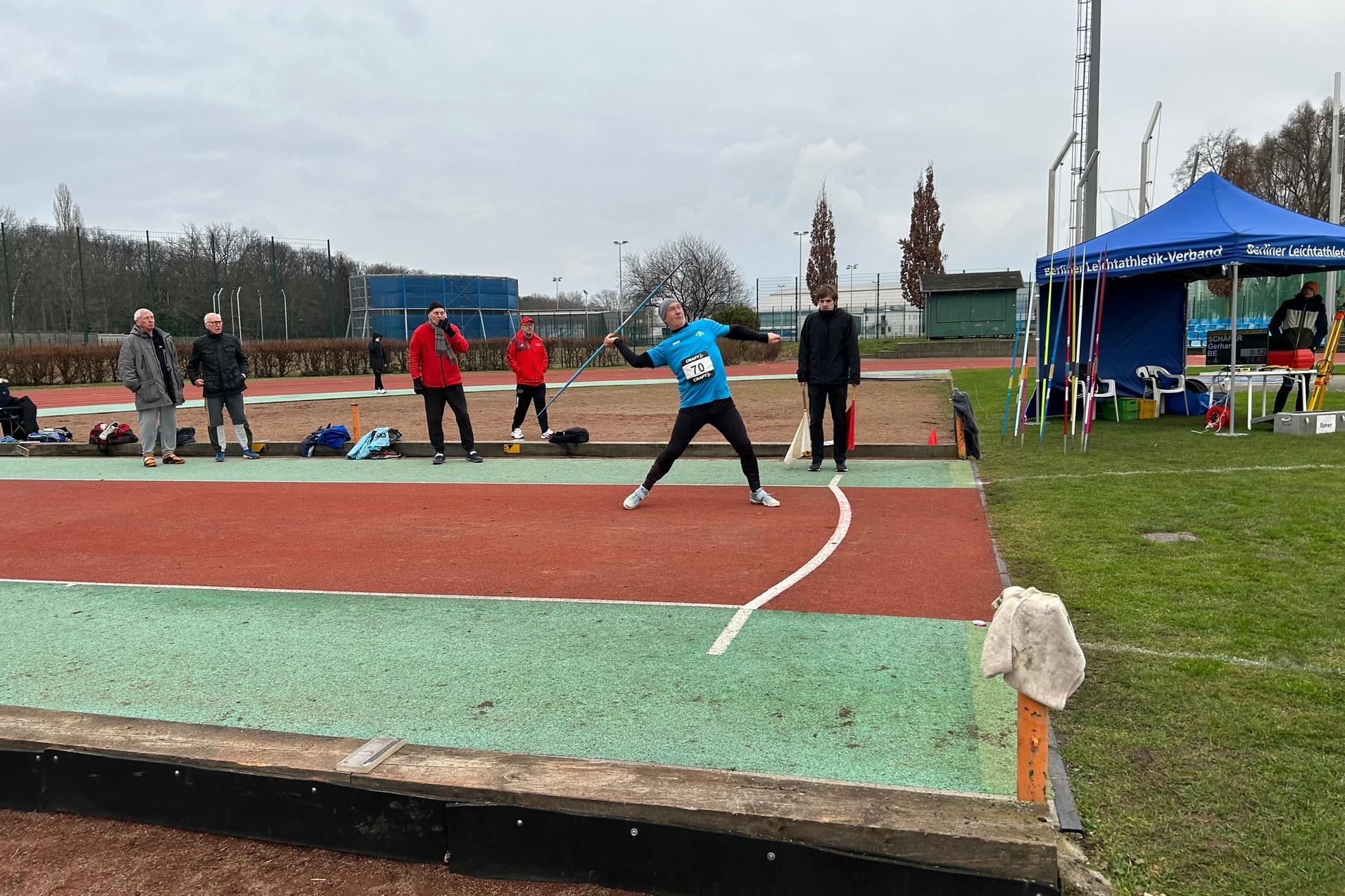 Athlet wirft eine Speer auf einem Wettkampfsportplatz, während Zuschauer und Trainer in der Nähe stehen.