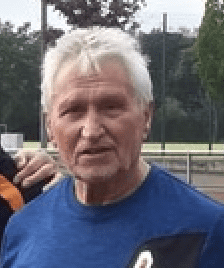Ein älterer Mann mit grauen Haaren und blauem Shirt steht im Freien, Hintergrund zeigt Sportanlagen und Bäume.
