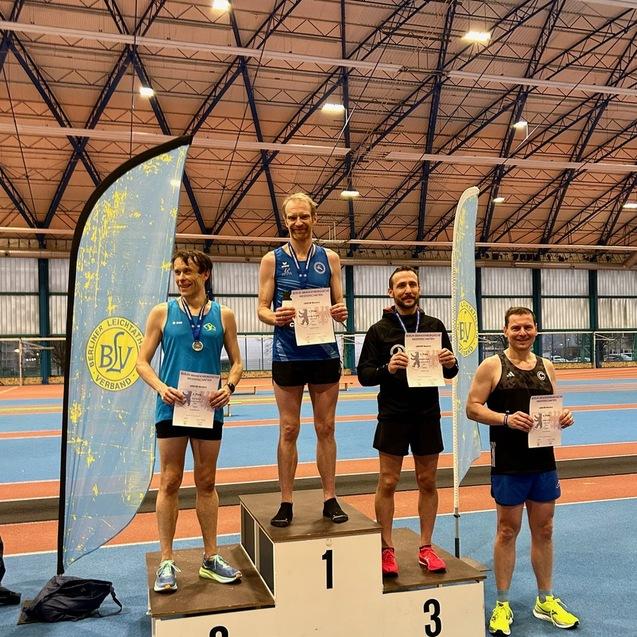 Vier m&auml;nnliche Athleten auf dem Siegertreppchen mit Urkunden in der Hand, im Hintergrund eine Sporthalle und ein Vereinsbanner.