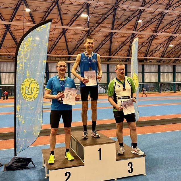 Drei hergestellte Sportler auf dem Siegertreppchen halten Urkunden; ein Banner des Berliner Leichtathletikverbands im Hintergrund.