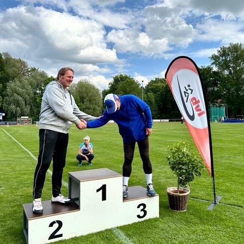 Zwei M&auml;nner auf einem Siegertreppchen in einem Stadion, w&auml;hrend ein Kind im Hintergrund sitzt und zuschaut.