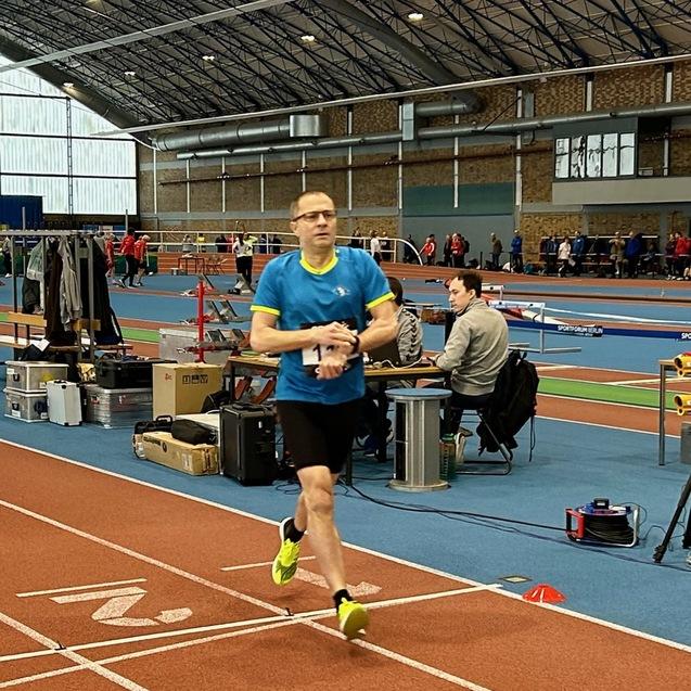 Ein Athlet in blauer Sportkleidung läuft auf einer Laufbahn in einer Sporthalle während eines Wettkampfs.